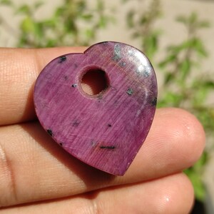 May include: A heart-shaped pendant made of pink ruby with a hole drilled through the top. The pendant has a smooth, polished surface and a dark pink colour.