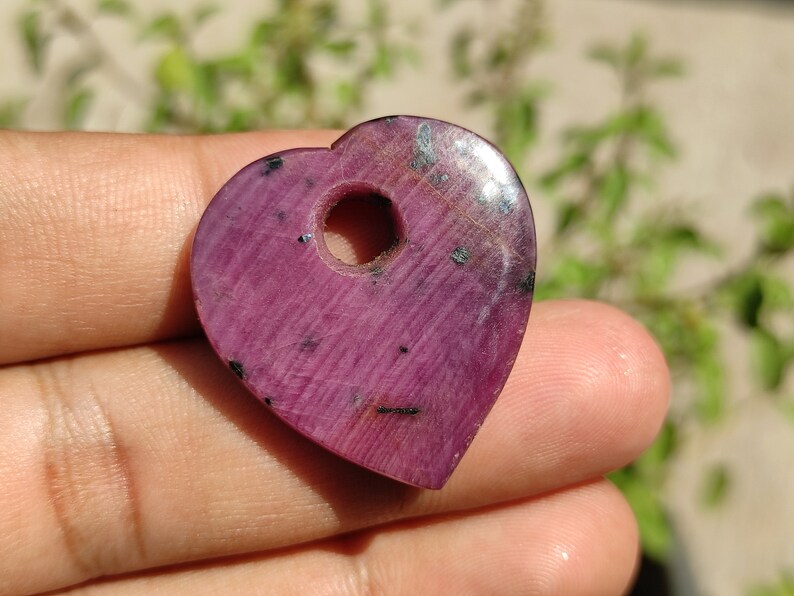 May include: A heart-shaped pendant made of pink ruby with a hole drilled through the top. The pendant has a smooth, polished surface and a dark pink colour.