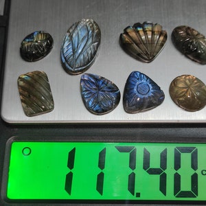 May include: A selection of carved labradorite gemstones in diverse shapes, such as ovals and teardrops, and a seashell design. The stones are on a digital scale, with a green display showing a weight of 17.40 ct.