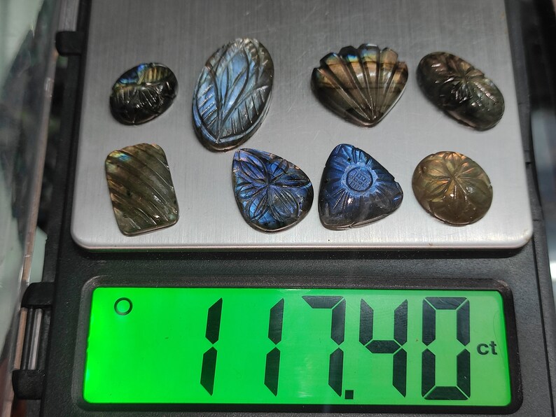 May include: A selection of carved labradorite gemstones in diverse shapes, such as ovals and teardrops, and a seashell design. The stones are on a digital scale, with a green display showing a weight of 17.40 ct.