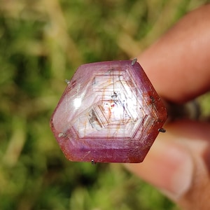 May include: A pink and red gemstone with a hexagonal shape and a star-like pattern in the center. The gemstone is set in a metal setting with prongs.