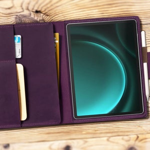 Może przedstawiać: Futroł folio wykonany z fioletowej skóry z wbudowanym gniazdem na rysik i kieszeniami na karty. Futerał jest otwarty, ukazując tablet z abstrakcyjnym niebiesko-zielonym tłem.