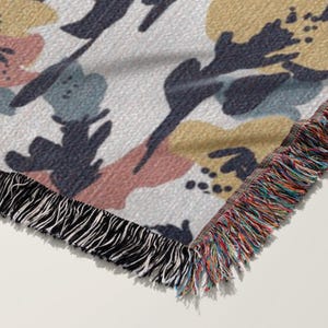 May include: A woven throw blanket with a floral pattern in muted colors. The design features abstract shapes in pink, blue, and yellow on a white background. The blanket has a fringed edge in black and a multi-colored fringe.