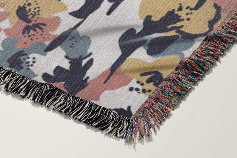 May include: A woven throw blanket with a floral pattern in muted colors. The design features abstract shapes in pink, blue, and yellow on a white background. The blanket has a fringed edge in black and a multi-colored fringe.