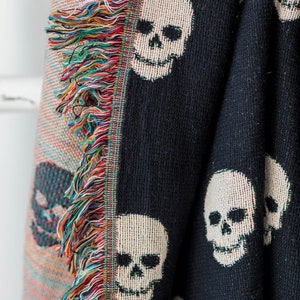 May include: Black woven throw blanket with a white skull pattern. The blanket has a fringe border with multicoloured threads.