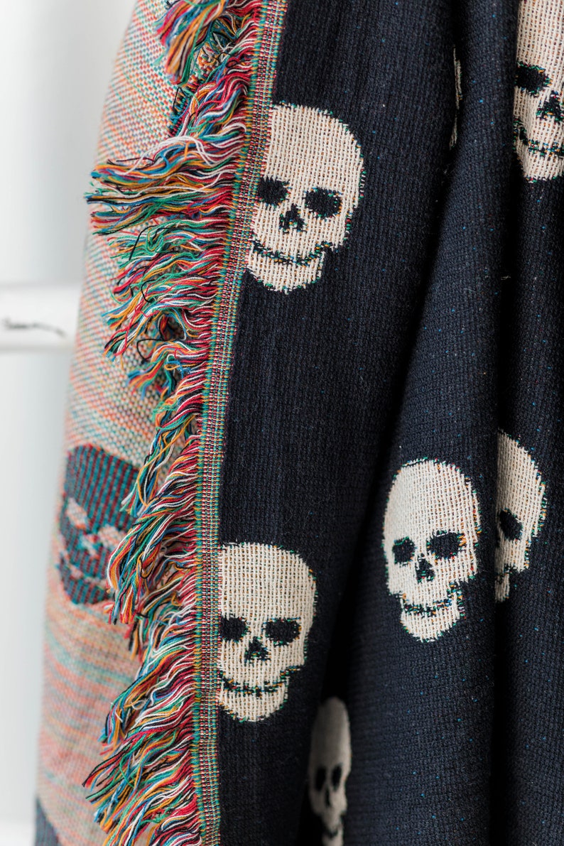 May include: Black woven throw blanket with a white skull pattern. The blanket has a fringe border with multicoloured threads.