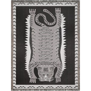 May include: A black and white woven tapestry featuring a stylized tiger design. The tiger is depicted upside down with its paws in the air and its tail curled around its body. The background is a solid black color.