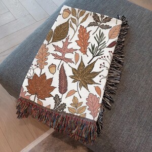 May include: A rectangular throw blanket with a white background and a repeating pattern of autumn leaves in shades of brown, orange, and green. The blanket has a fringed edge with multiple colors.