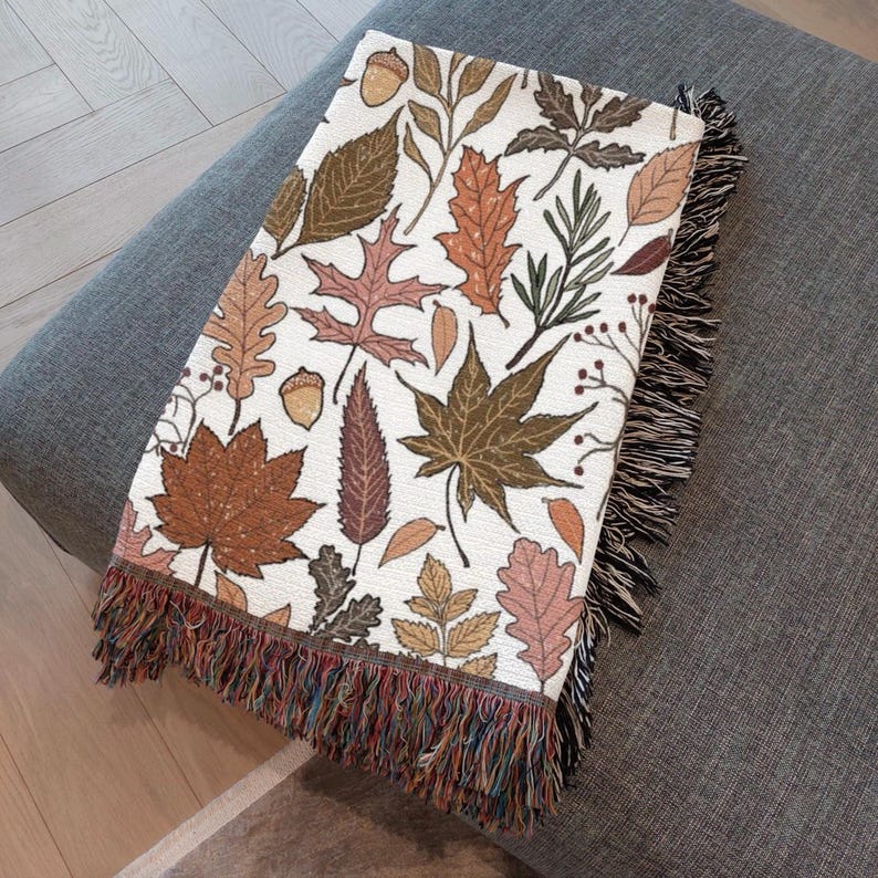 May include: A rectangular throw blanket with a white background and a repeating pattern of autumn leaves in shades of brown, orange, and green. The blanket has a fringed edge with multiple colors.