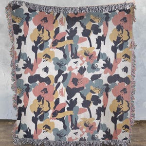 May include: A woven throw blanket with a floral pattern. The design features large, stylized flowers in shades of pink, yellow, blue, and gray on a cream background. The blanket has a fringed edge.