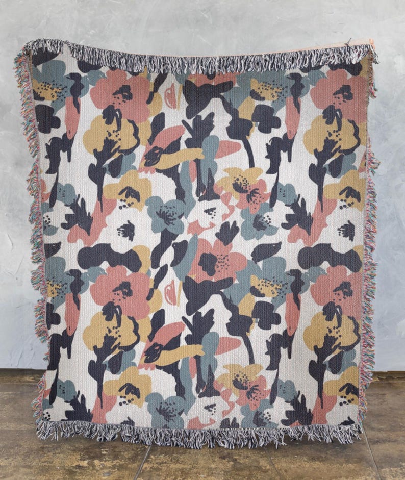 May include: A woven throw blanket with a floral pattern. The design features large, stylized flowers in shades of pink, yellow, blue, and gray on a cream background. The blanket has a fringed edge.