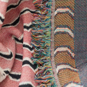 May include: A pink and black striped woven throw blanket with a fringe border. The fringe is made of multi-colored yarn.
