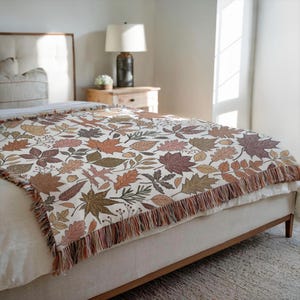 May include: A woven throw blanket featuring a repeating pattern of autumn leaves in shades of brown, orange, and green on a cream background. The blanket has a fringed edge and is draped over a bed.