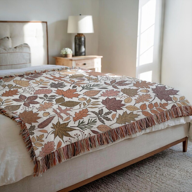 May include: A woven throw blanket featuring a repeating pattern of autumn leaves in shades of brown, orange, and green on a cream background. The blanket has a fringed edge and is draped over a bed.