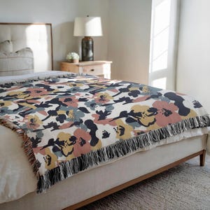 May include: A floral patterned throw blanket in shades of pink, yellow, blue, and navy on a cream background. The blanket has a fringed edge and is draped over a bed. The design features large, abstract flowers.