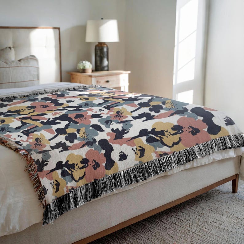 May include: A floral patterned throw blanket in shades of pink, yellow, blue, and navy on a cream background. The blanket has a fringed edge and is draped over a bed. The design features large, abstract flowers.
