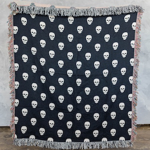 May include: Black woven throw blanket with a white skull pattern. The blanket has fringe on all sides.