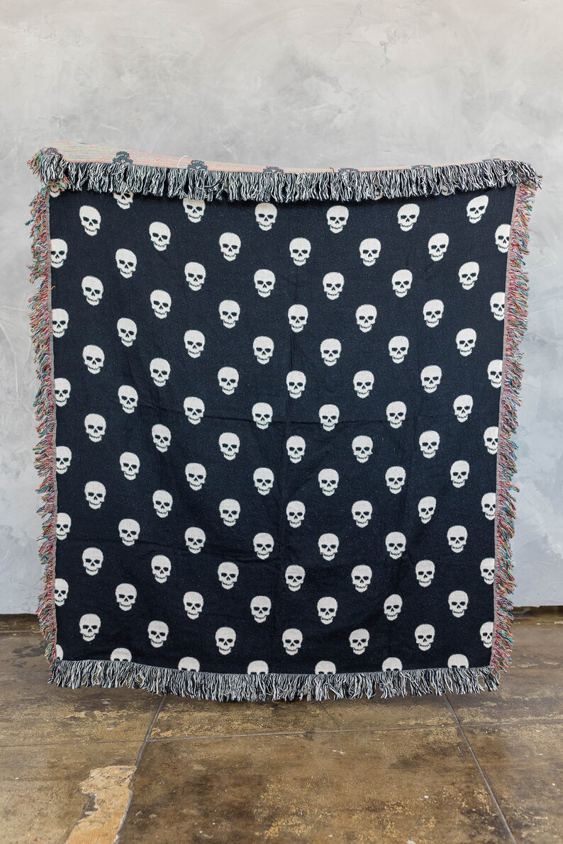 May include: Black woven throw blanket with a white skull pattern. The blanket has fringe on all sides.