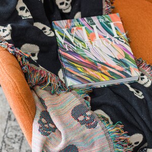May include: A black and white throw blanket with a skull pattern and colourful fringe is draped over an orange armchair. A book titled "Wild Interiors" by Hilton Carter is on top of the blanket. The book cover features a colourful abstract design with the text "Beautiful Plants in Beautiful Spaces."