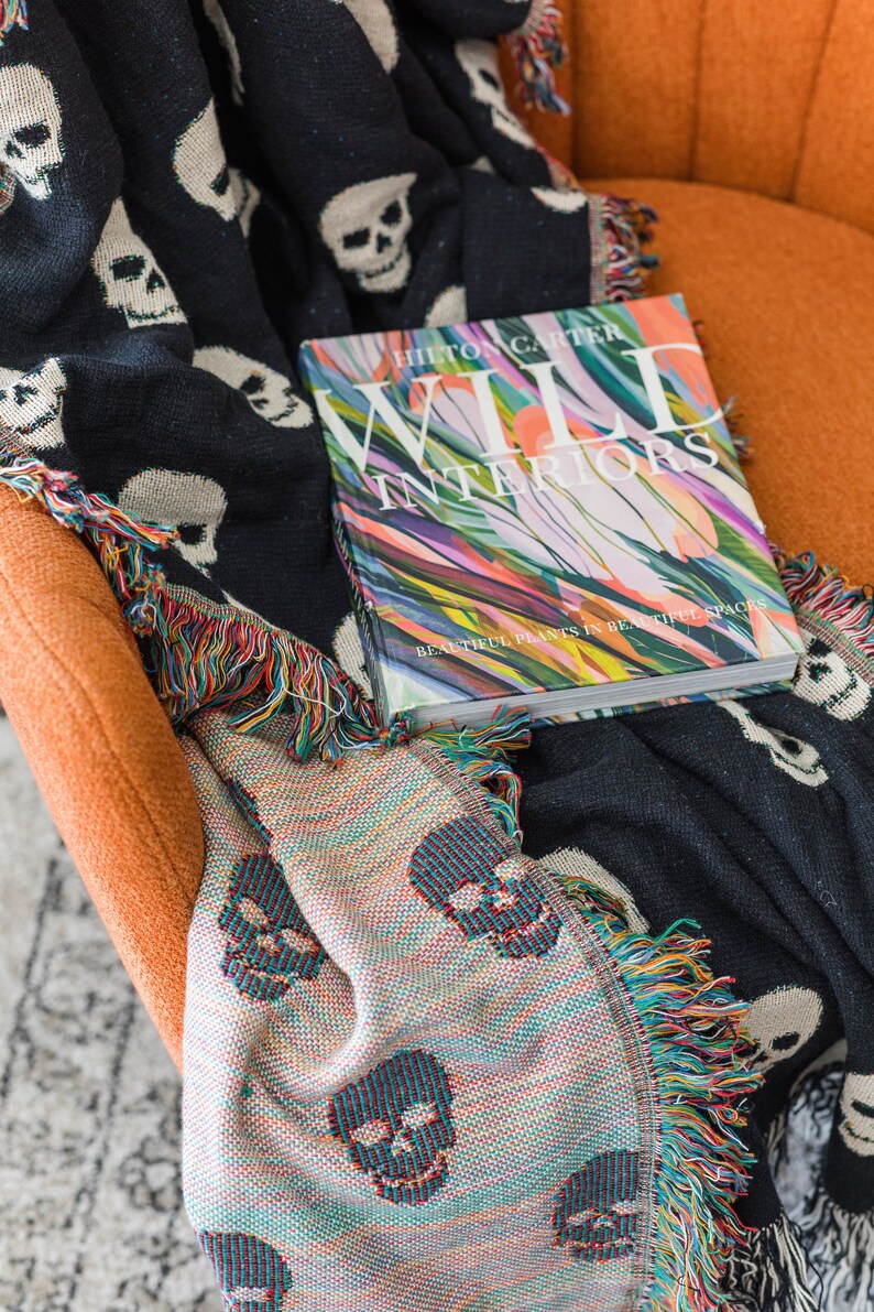 May include: A black and white throw blanket with a skull pattern and colourful fringe is draped over an orange armchair. A book titled "Wild Interiors" by Hilton Carter is on top of the blanket. The book cover features a colourful abstract design with the text "Beautiful Plants in Beautiful Spaces."