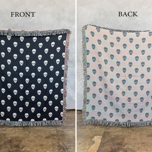 May include: A black and white woven throw blanket with a skull pattern on the front and a pink and blue striped pattern with a skull pattern on the back. The blanket has fringe on all sides.