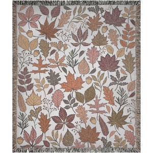 May include: Woven throw blanket featuring a repeating pattern of autumn leaves and acorns in shades of brown, orange, and pink on a cream background. The blanket has a fringed edge and is suitable for home decor.