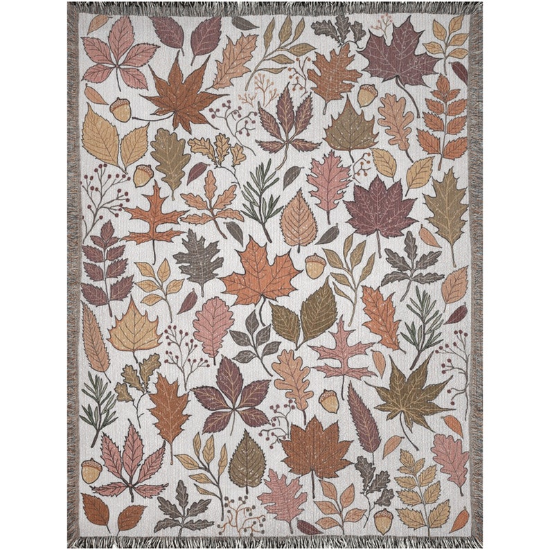 May include: A woven throw blanket featuring a repeating pattern of autumn leaves in shades of brown, orange, and pink on a cream background. The blanket has a fringed edge and is suitable for home decor.