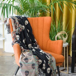 May include: A black and white skull patterned throw blanket draped over an orange armchair. The blanket has a fringe border and is made of woven fabric.