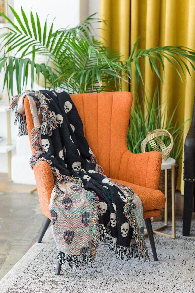 May include: A black and white skull patterned throw blanket draped over an orange armchair. The blanket has a fringe border and is made of woven fabric.