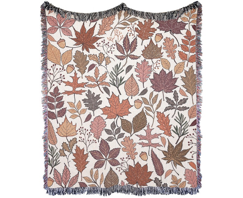 May include: A woven throw blanket featuring a repeating pattern of autumn leaves and acorns in shades of brown, orange, and pink on a cream background. The blanket has a fringed edge.