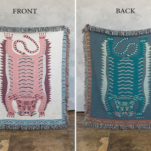 May include: A woven tapestry throw blanket features a pink and teal tiger design. The front of the blanket features a pink tiger with black stripes, while the back features a teal tiger with black stripes. The blanket has a fringe border.