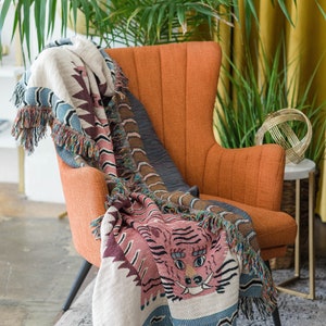 May include: A woven throw blanket with a pink tiger face design and blue and brown accents. The blanket is draped over an orange armchair in a living room setting.