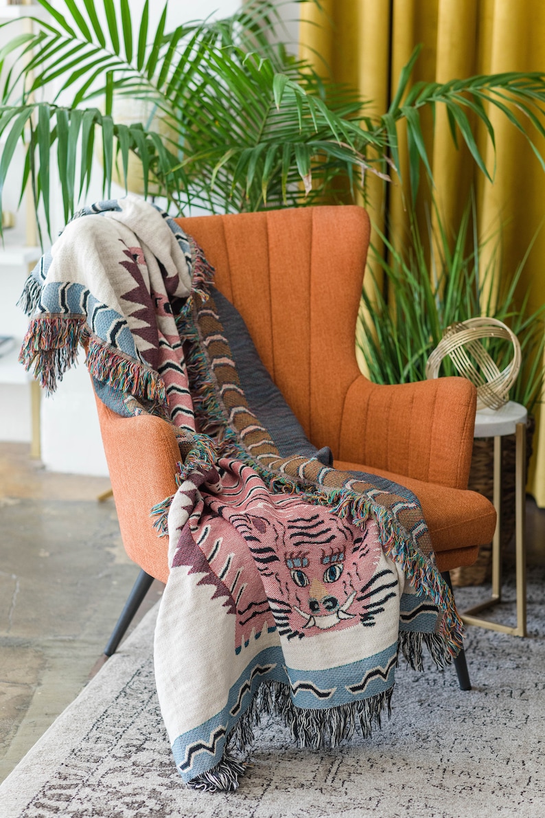 May include: A woven throw blanket with a pink tiger face design and blue and brown accents. The blanket is draped over an orange armchair in a living room setting.