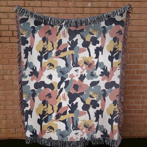 May include: A woven throw blanket with a floral pattern in shades of pink, yellow, blue, and navy on a white background. The blanket has a fringed edge and is displayed against a brick wall.
