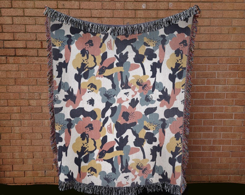 May include: A woven throw blanket with a floral pattern in shades of pink, yellow, blue, and navy on a white background. The blanket has a fringed edge and is displayed against a brick wall.