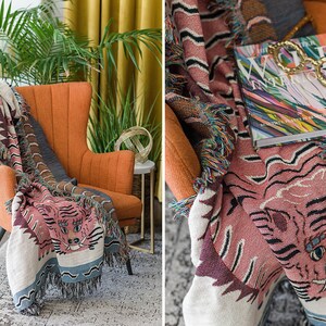 May include: A pink and white woven throw blanket with a tiger face design. The blanket is draped over an orange armchair with a book and glasses on top. The book cover features a colorful abstract design and the text "The World Interiors".