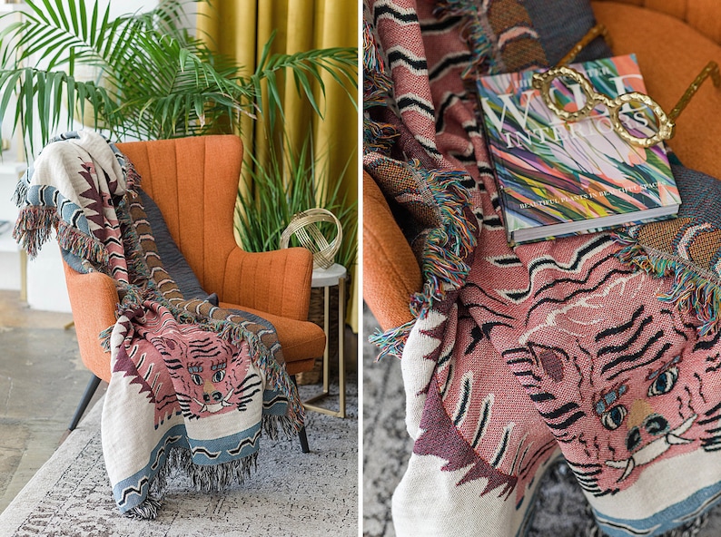 May include: A pink and white woven throw blanket with a tiger face design. The blanket is draped over an orange armchair with a book and glasses on top. The book cover features a colorful abstract design and the text "The World Interiors".