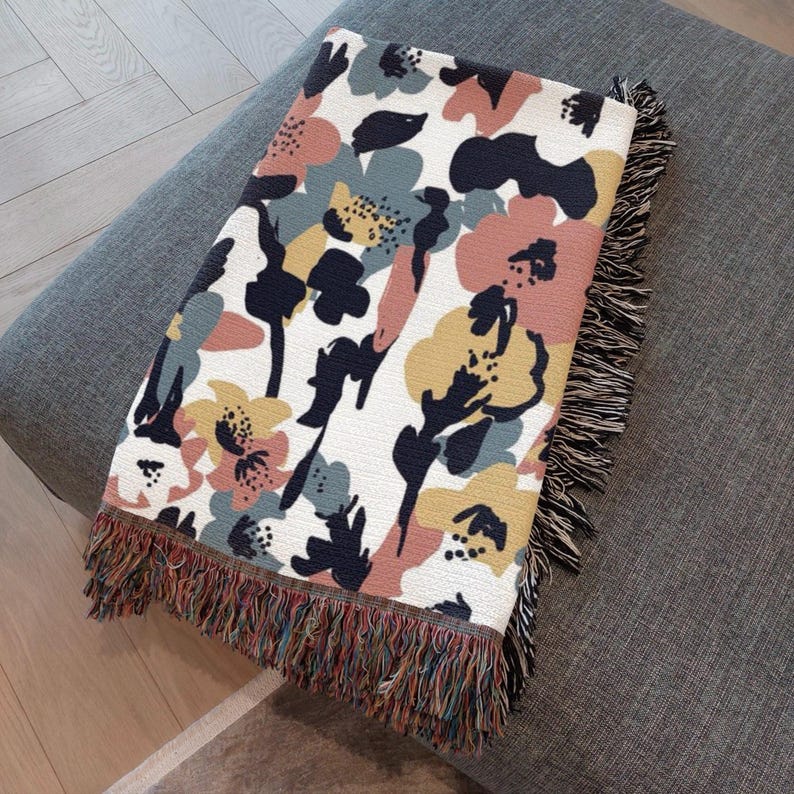May include: A floral patterned throw blanket with a white background, featuring pink, yellow, blue, and black flower designs. The blanket has a fringed edge with multiple colors. The blanket is folded on a gray surface.