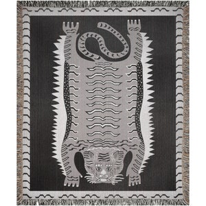 May include: A woven tapestry featuring a gray tiger with black stripes, lying on its back with its paws in the air. The tiger is on a black background with a white border.