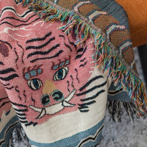 May include: A pink and white woven throw blanket with a tiger face design. The blanket has a fringe border with multi-colored threads.