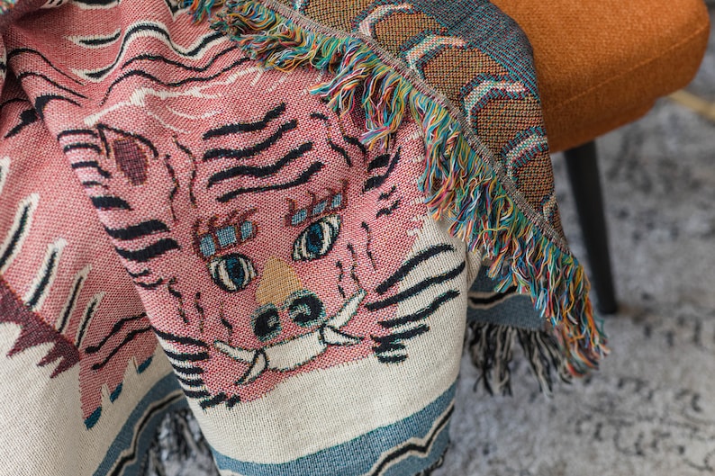 May include: A pink and white woven throw blanket with a tiger face design. The blanket has a fringe border with multi-colored threads.