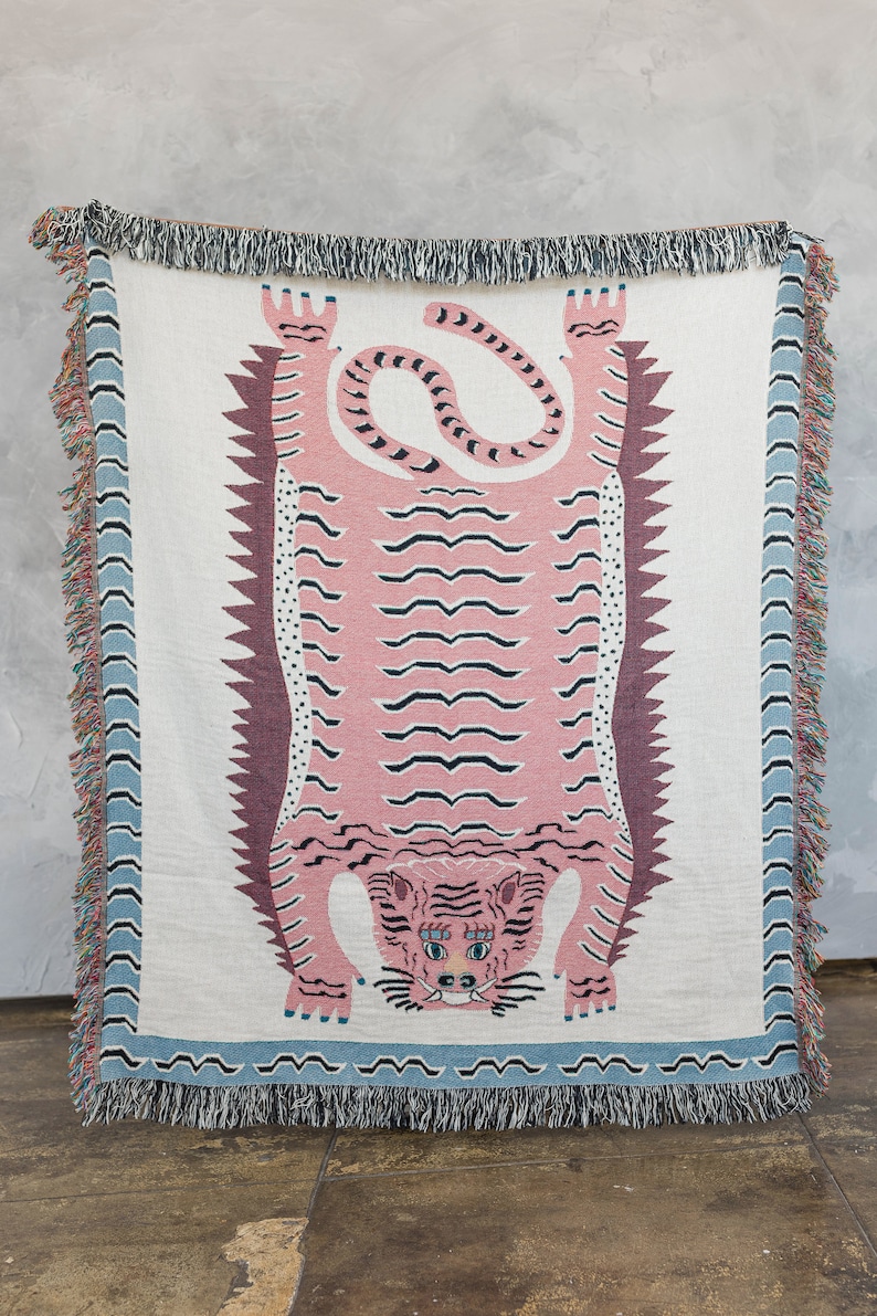 May include: A pink and white woven throw blanket with a pink tiger design. The tiger is upside down with its paws in the air. The blanket has a blue and white fringe border.