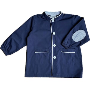 May include: A navy blue long-sleeve smock featuring a mandarin collar, white buttons, and two front pockets. The collar and elbow patches are accented with a white and navy blue pinstripe pattern. A white label with text is inside the collar.