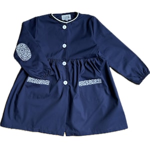 Peut inclure: Une blouse bleu marine à manches longues avec un col rond bordé de blanc, quatre boutons blancs sur le devant et deux poches plaquées à bords fleuris. Un écusson ovale à motif floral est visible sur le coude droit de la manche.