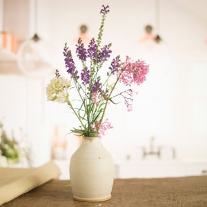 May include: A white ceramic vase with a textured surface holds a bouquet of purple, pink, and white flowers. The vase is sitting on a wooden table.