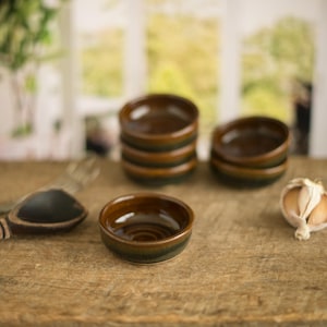 May include: A collection of small, brown ceramic bowls with a green rim, stacked and arranged on a wooden surface. A wooden spoon and a garlic bulb are also present, suggesting a culinary theme.