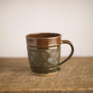 May include: A handmade ceramic mug with a brown glazed rim and handle. The body of the mug is a darker green-brown with a wave pattern. The mug sits on a wooden surface.