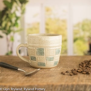Peut inclure: Une tasse en céramique blanche avec un motif à carreaux bleu clair et beige. La tasse a un glaçage moucheté et une poignée arrondie.