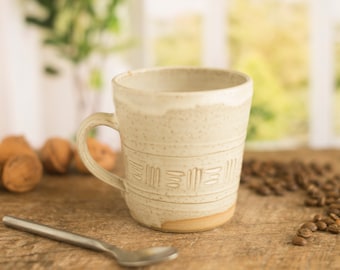May include: A white ceramic mug with a textured design and a brown handle. The mug is sitting on a wooden surface with coffee beans scattered around it.