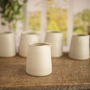 Op de afbeelding: Vijf witte keramische roomkannen met een gespikkeld glazuur. De kannen staan in een rij op een houten oppervlak.